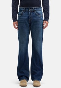 Dżinsy denim w ciemnym odcieniu, szerokie nogawki, styl z pięcioma kieszeniami. Posiadają zapięcie na przedni guzik i delikatne przetarcia na nogawkach. Widoczne zamszowe buty.