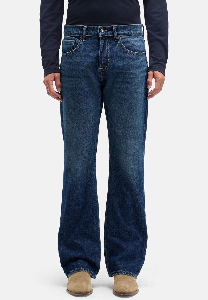 Dżinsy denim w ciemnym odcieniu, szerokie nogawki, styl z pięcioma kieszeniami. Posiadają zapięcie na przedni guzik i delikatne przetarcia na nogawkach. Widoczne zamszowe buty.