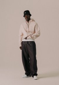 Haut de survêtement oversize rose clair avec cordons, pantalon ample noir et baskets blanches. Un chapeau seau noir et des lunettes complètent le look.