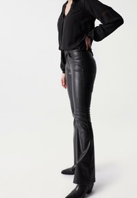 Pantaloni in pelle nera svasati con una texture liscia, vita alta e dettagli di cucitura sottili. Abbinati a una blusa nera a maniche lunghe.