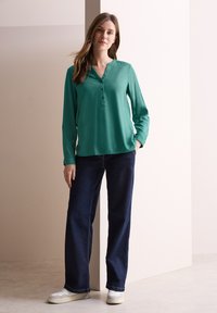 Blusa lunga teal con colletto a punta e pattina con bottoni abbinata a jeans a gamba larga blu scuro e sneaker bianche. Tessuto di una consistenza liscia.