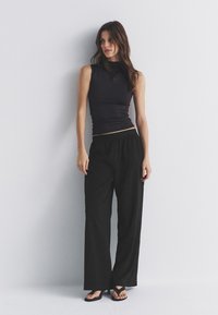 Haut noir moulant sans manches associé à un pantalon noir ample avec taille élastique. Modèle portant des tongs noires. Fond simple et neutre.