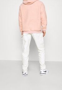 Lichtroze, fluffy fleecejack met capuchon, gecombineerd met witte cargobroeken. Witte sneakers met zwarte accenten. Casual achterkant.