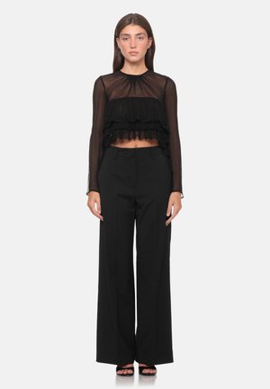 Donna che indossa un crop top nero trasparente a maniche lunghe con volant, pantaloni neri a gamba larga e vita alta, e tacchi neri aperti, in piedi su sfondo bianco.