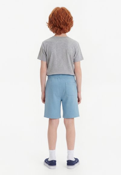 Παιδί με σγουρά κόκκινα μαλλιά στέκεται με την πλάτη γυρισμένη, φοράει γκρι μπλουζάκι, ανοιχτό μπλε σορτς, άσπρες κάλτσες και μπλε σκούρα αθλητικά παπούτσια σε λευκό φόντο.