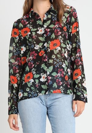 Kobieta w czarnej przezroczystej kwiatowej bluzce z czerwonymi, białymi i fioletowymi kwiatami, w połączeniu z jasnoniebieskimi dżinsami.