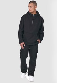 Sudadera negra con cremallera y bolsillo canguro, decorada con un logotipo bordado, combinada con unos pantalones cargo a juego que cuentan con bolsillos laterales, llevados con zapatillas blancas.