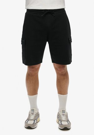 Pantalones cortos cargo negros de tela suave, con dos bolsillos laterales y una cintura con cordón ajustable. Combinados con calcetines de color claro y zapatillas deportivas.