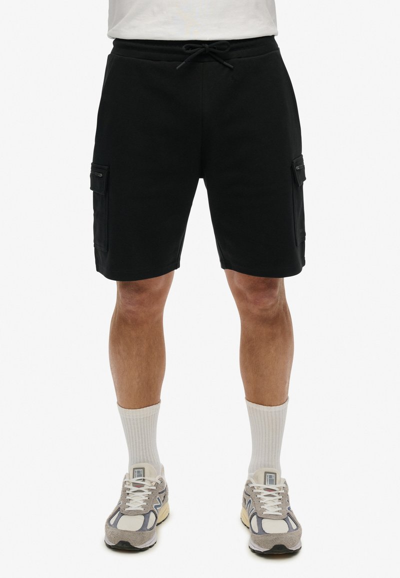 Czarne szorty cargo wykonane z miękkiego materiału, z dwoma kieszeniami bocznymi i regulowanym paskiem. W komplecie z jasnymi skarpetkami i trampkami.