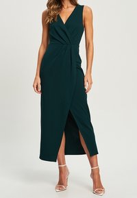 Vestido maxi sin mangas de color verde oscuro con escote en V, detalle de frente retorcido y una abertura lateral. Tejido suave, de largo hasta el tobillo, combinado con tacones de tiras.