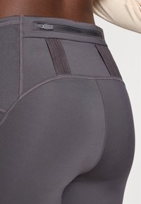 Høyt liv, tettsittende leggings i mørkegrått stoff med en glatt tekstur. Har en glidelåslomme og subtile sømdetaljer i livet.
