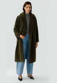 Olivgrüner, strukturierter Mantel mit Schalkragen, kombiniert mit einem grauen Pullover und hellblauen, weit geschnittenen Jeans. Schwarze spitz zulaufende Schuhe.