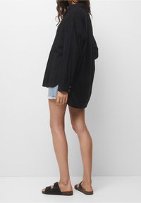 Chemise noire à manches longues avec col, coupe décontractée et manches à boutons, associée à un short en denim usé et des sandales noires.