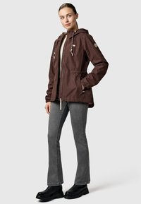 Femme portant une veste brune zippée à capuche, un jean évasé gris et des bottes noires, debout avec une main dans la poche de la veste.