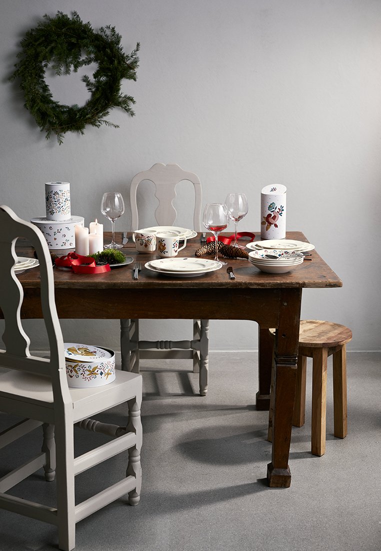 Drewniany stół jadalny z czterema krzesłami, porcelanowymi naczyniami, szkłem, białymi świecami oraz dekoracyjnymi pudełkami z kwiatowymi wzorami.