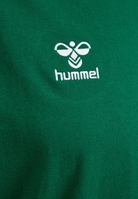 Groene t-shirt met een geborduurd wit logo met een gestileerde bij en de merknaam "hummel" in kleine letters. Glad textuur.