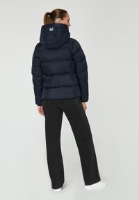 Marineblauer Pufferjacke mit Kapuze, ausgestattet mit gesteppte Abschnitten und einem Logo auf der Rückseite. Kombiniert mit schwarzen Jeans und weißen Schuhen.