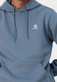 Sudadera azul hecha de una tela suave, con un bolsillo canguro, cordones y un pequeño logo blanco de Converse en el pecho.