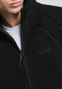 Jachetă din fleece neagră cu guler înalt, închidere cu fermoar și logo embosat subtil. Prezintă un material texturat și fermoare contrastante.