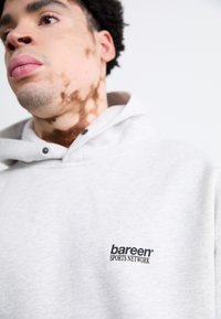 Lys grå hættejakke lavet af blødt stof, der har et logo foran, en justerbar hætte og sorte trykknapper på kraven.