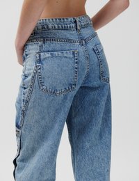 Άτομο που φοράει τζιν παντελόνι με ψηλή μέση σε ανοιχτό γαλάζιο χρώμα, με ορατές πίσω τσέπες και λεπτομέρειες με κουμπιά στο πλάι, σε ουδέτερο φόντο.