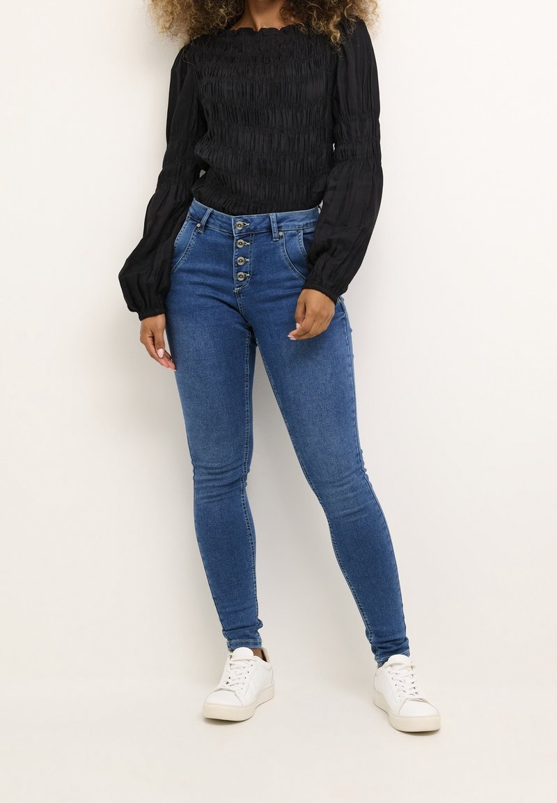 Jeans de mezclilla azul de talle alto con cierre frontal de cinco botones, corte ajustado y un ligero estiramiento. Combinados con una blusa negra de manga larga con fruncido.