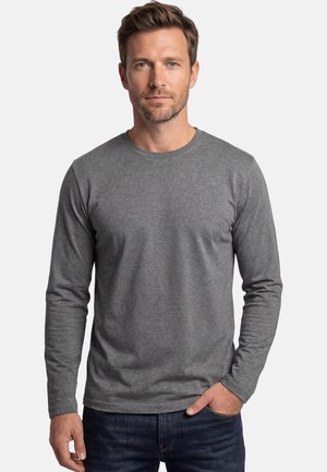 Uomo con capelli castani corti e barba, che indossa una maglia grigia a maniche lunghe semplice e jeans scuri, in piedi con una mano in tasca.