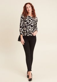 Langärmlige Bluse mit schwarzem und weißem Blumenmuster, elastischem Bund, kombiniert mit figurbetonten schwarzen Jeans und schwarzen High Heels.