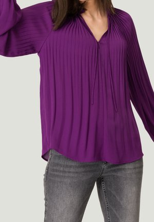 Femme portant un chemisier plissé violet à manches longues avec un col en V et un détail à nouer, associé à un jean gris délavé sur un fond uni.