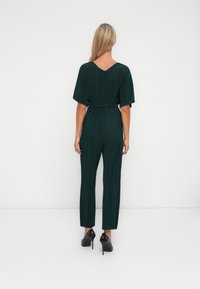 Kvinne med langt blondt hår iført mørk grønn plissé jumpsuit med belte og svarte høye heler, står mot en enkel hvit bakgrunn.