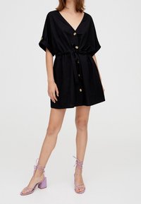 Czarna sukienka zapinana na guziki z dekoltem w serek i krótkimi podwiniętymi rękawami. Posiada taliowanie i złote guziki. Połączona z lawendowymi sandałkami na obcasie z paskami.