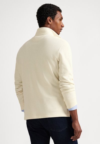 Muž s krátkými kudrnatými vlasy, oblečený v krémovém svetru s dlouhým rukávem a tmavých džínách, stojící bokem na neutrálním pozadí.