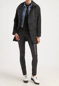 Textured zwarte jas met zilveren rits, spijkerjack eronder, gecombineerd met glanzende zwarte broek en witte schoenen met zwarte accenten.