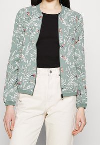 Zip-front bomberjacka i dämpad grön med blomm- och fågelmönster. Ribbstickade muddar och nederdel, tillsammans med ett lätt, strukturerat tyg.