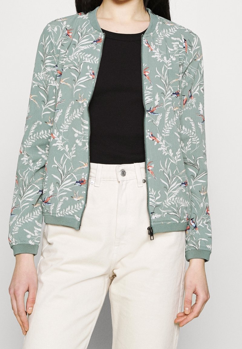 Zip-front bomberjacka i dämpad grön med blomm- och fågelmönster. Ribbstickade muddar och nederdel, tillsammans med ett lätt, strukturerat tyg.