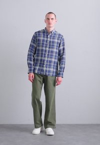 Πουκάμισο με κουμπιά σε μπλε καρώ ύφασμα; πράσινα παντελόνια; λευκά αθλητικά παπούτσια. Το μοντέλο στέκεται μπροστά σε απλό γκρι φόντο, αναδεικνύοντας τις λεπτομέρειες του ρούχου.