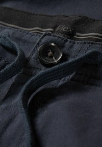 Närbild av mörkblå byxor med en svart knapp och synlig dragsko, som visar midjebandsetiketten med texten "Flex the go".