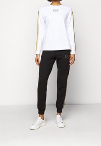 Felpa bianca con strisce dorate scintillanti e pantaloni neri abbinati con dettagli dorati e un logo. Sneakers bianche completano l'outfit.