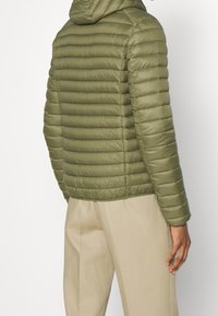 Giacca piumino isolante verde oliva con cuciture orizzontali, cappuccio e polsini elasticizzati. Indossata con pantaloni di colore chiaro.