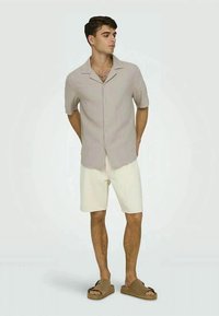 Camicia a maniche corte di colore beige chiaro con colletto, abbinata a pantaloni corti bianchi e sandali scivolosi marroni. La camicia ha un motivo testurizzato.