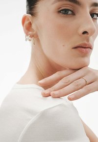 Pendientes de aro de oro y un delicado anillo de oro adornan una mano que reposa sobre un cuello. La blusa es blanca con una textura acanalada. La piel parece suave.