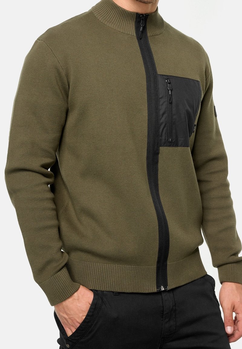 Homme portant un pull zippé vert olive avec une poche poitrine zippée noire et un pantalon noir, une main dans la poche.