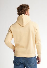 Felpa gialla chiara con una texture morbida e cappuccio regolabile, abbinata a jeans blu. La vista posteriore mostra una vestibilità rilassata e cuciture pulite.