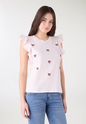 Giovane donna che indossa una maglietta senza maniche color rosa chiaro con volant e piccoli motivi di ciliegie rosse, abbinata a jeans blu, su sfondo chiaro.