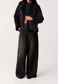 Schwarze Fake-Schafslederjacke mit Reißverschluss und breiten Revers, kombiniert mit locker sitzenden schwarzen Jeans und Stiefeletten.