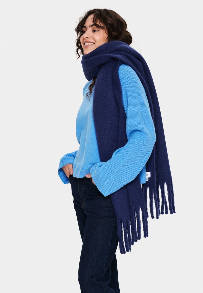 Blue cashmere scarf with long fringes draped around the neck, paired with a light blue sweater and dark jeans.