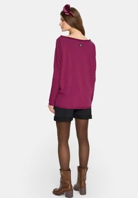 Magienta bluzka z długim rękawem o szerokim dekolcie, metka z tyłu, połączona z czarnymi szortami i brązowymi botkami za kostkę. Brązowa opaska na głowę z kwiatuszkowym detalem.