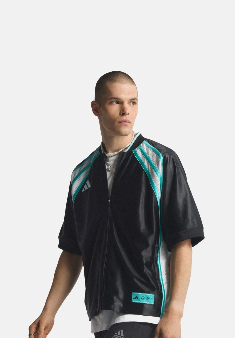 Junger Mann mit kurzem Haarschnitt, der eine schwarze und türkisfarbene Adidas-Sportjacke über einem weißen Hemd trägt und seitlich vor einem schlichten Hintergrund blickt.