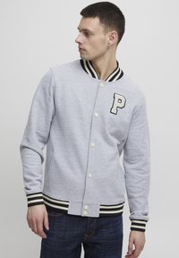 Grå varsityjacket med svart og kremstripete krage og mansjetter, med et «P»-merke og trykknapper. Glatt tekstur.