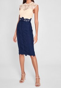 Krajkové šaty s béžovým krajkovým topem a námořnicky modrou krajkovou sukní s květinovými vzory a siluetou přesýpacích hodin. Look doplňují stiletto podpatky.